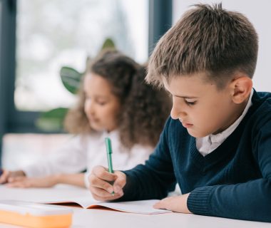 Concentrated,Schoolboy,Sitting,At,Desk,And,Writing,In,Exercise,Book